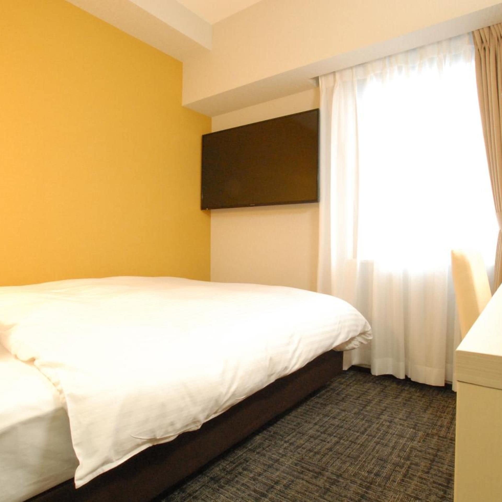 A hotel room with a bed, a flat-screen TV mounted on the wall, and a window with curtains.