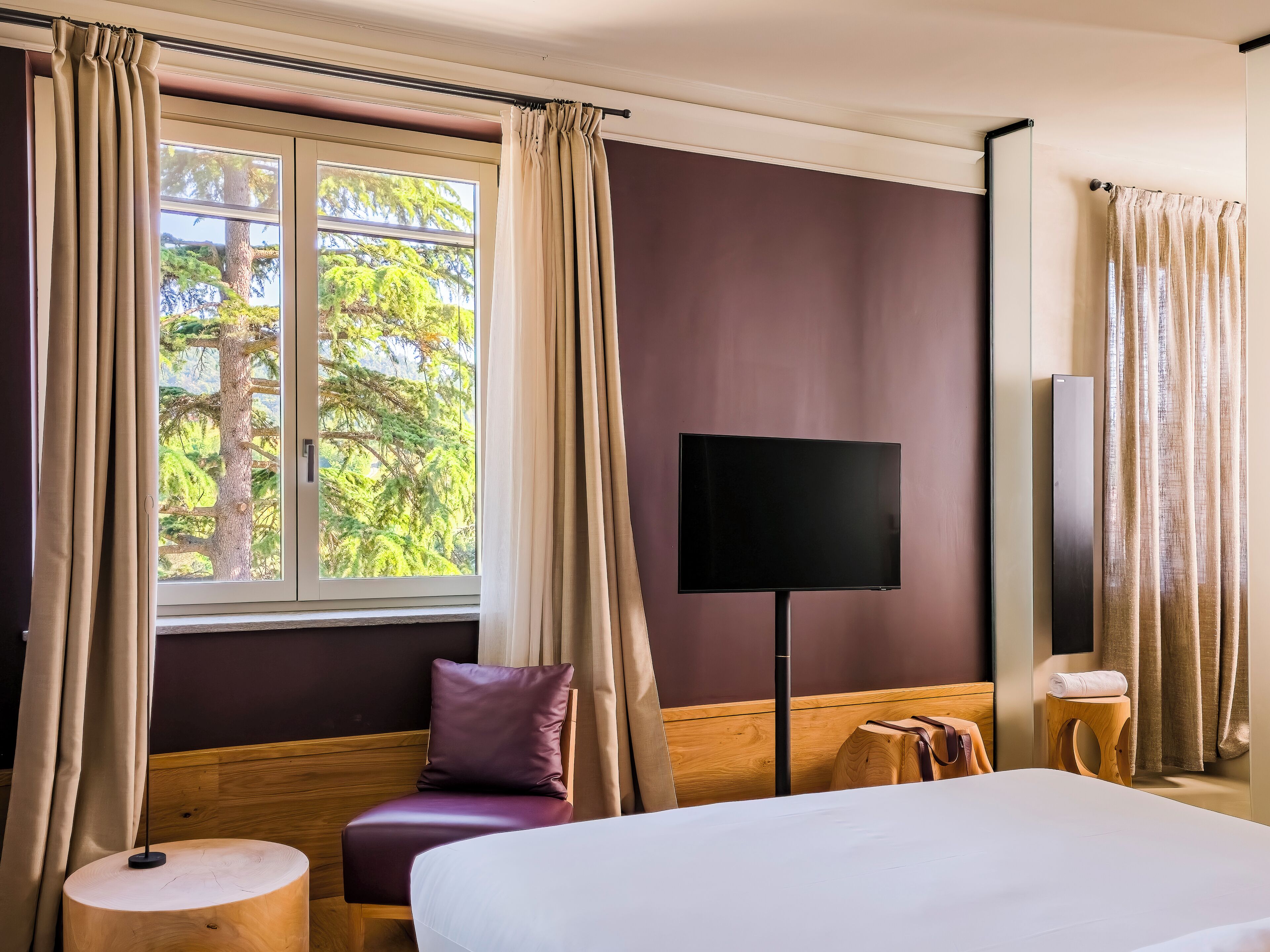 Une chambre d’hôtel avec un lit, une télévision, une fenêtre avec des rideaux, une chaise en bois et une petite table ronde.