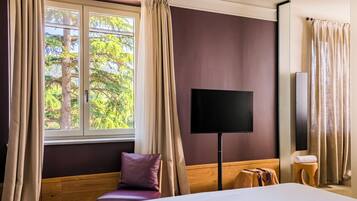 Une chambre d’hôtel avec un lit, une télévision, une fenêtre avec des rideaux, une chaise en bois et une petite table ronde.