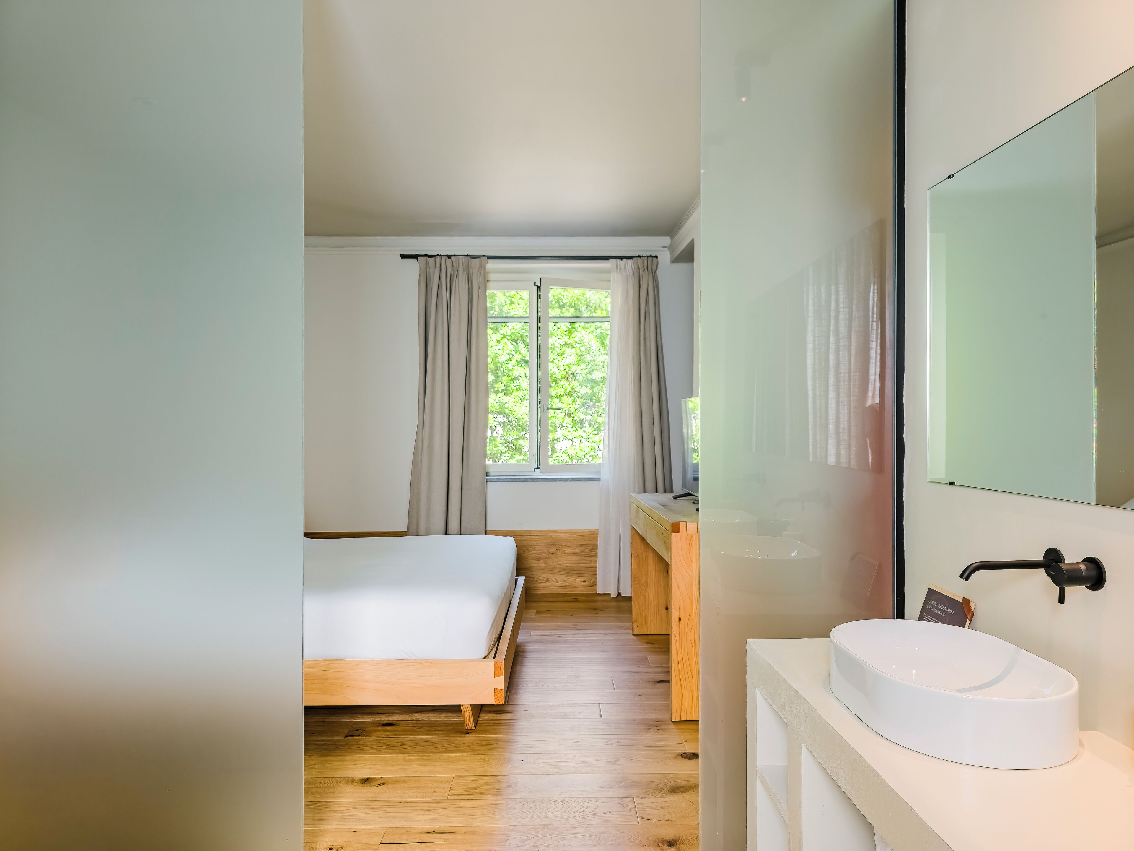 A bedroom with a wooden bed, a white bedside table, a large window with curtains, and a bathroom with a white sink and mirror.