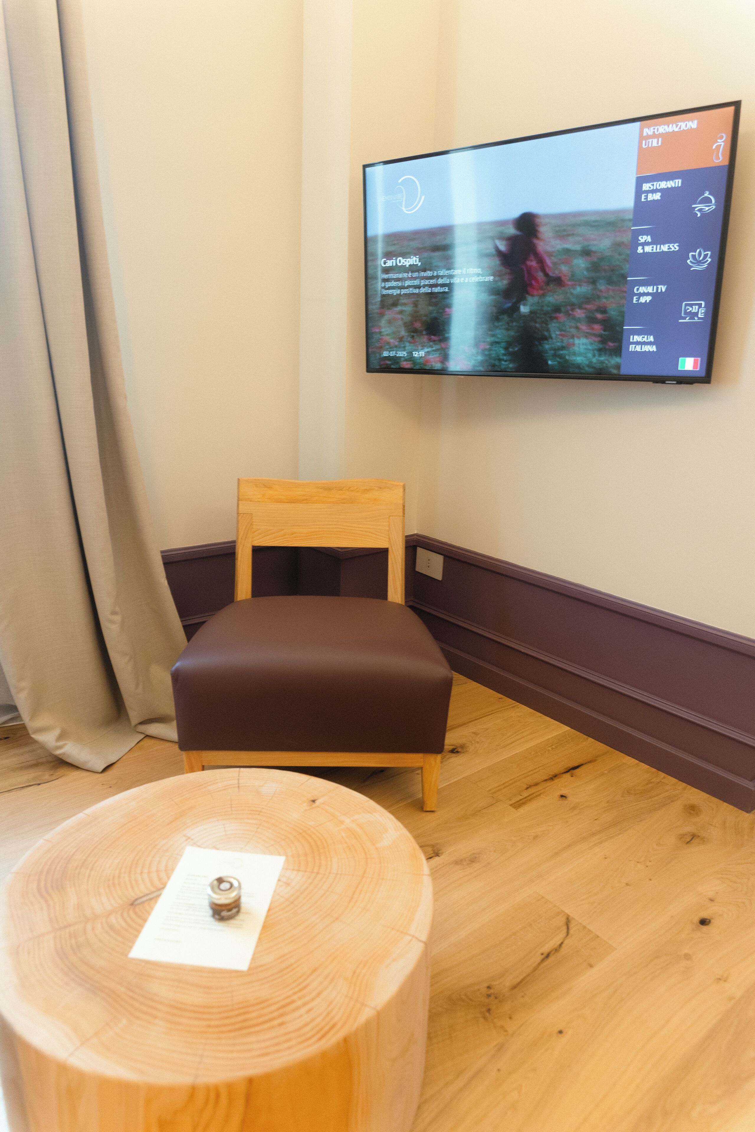 A room with a wooden chair, a round wooden table, and a wall-mounted TV displaying a menu.