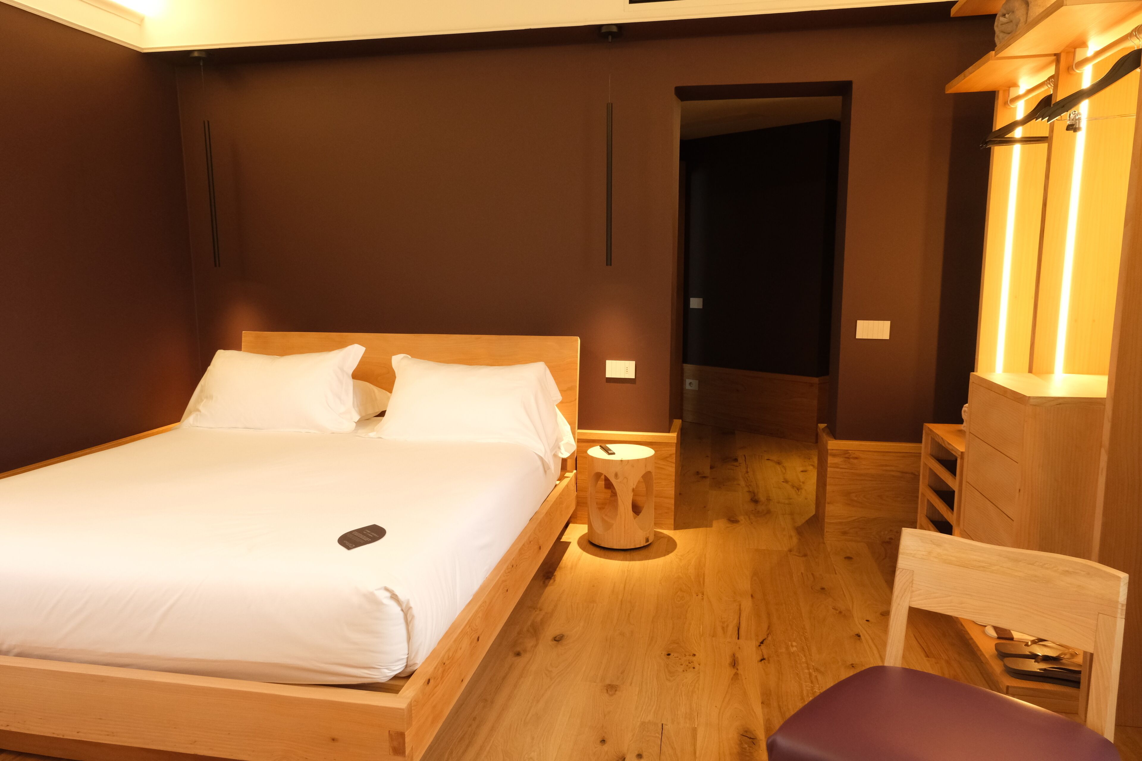 Ein hölzernes Bett mit weißen Leinen, ein Nachttisch und ein Stuhl in einem Zimmer mit Holzboden und -wänden.