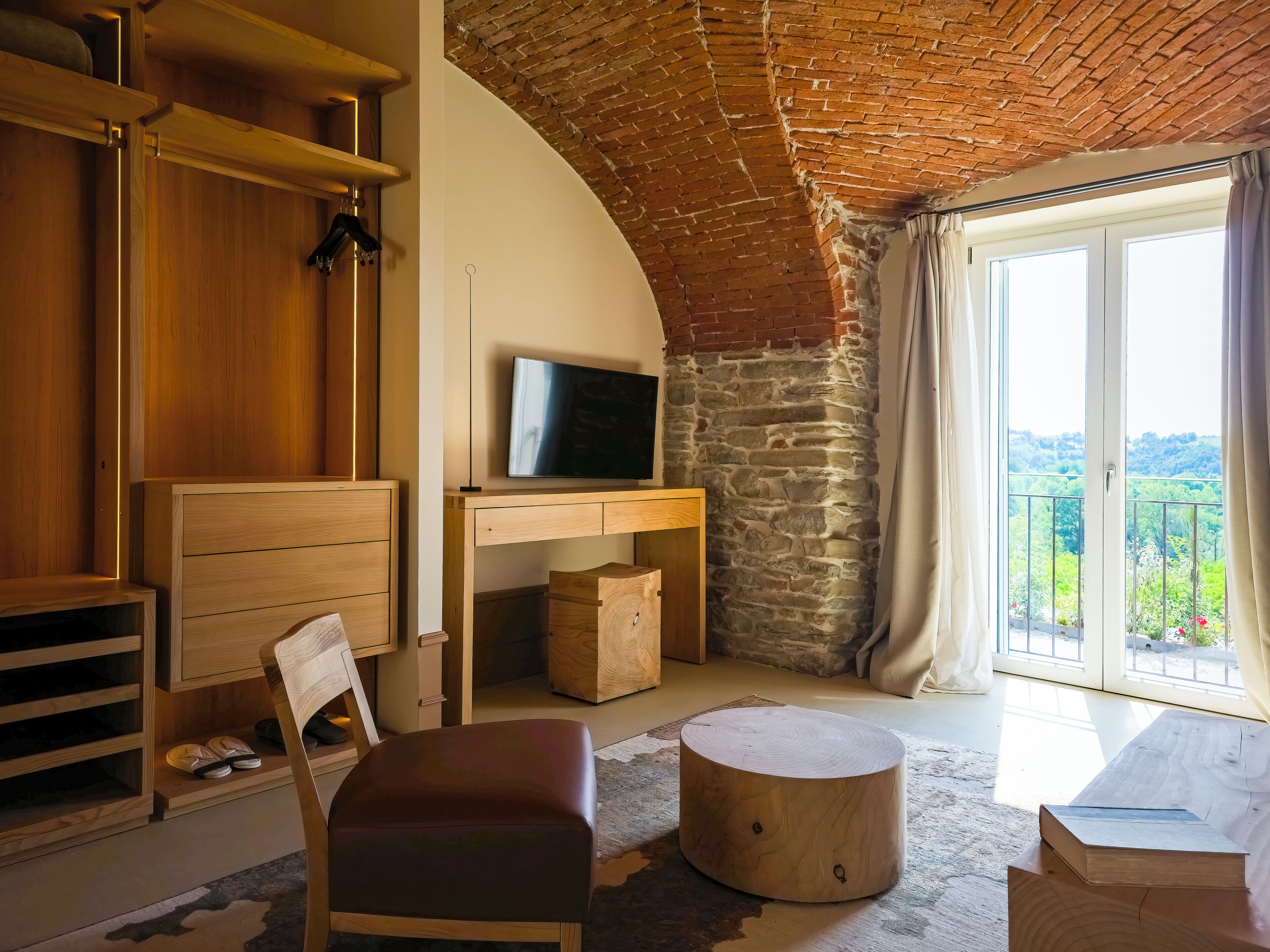 A cozy room with a wooden desk, a television, a comfortable chair, and a large window with a view of greenery.