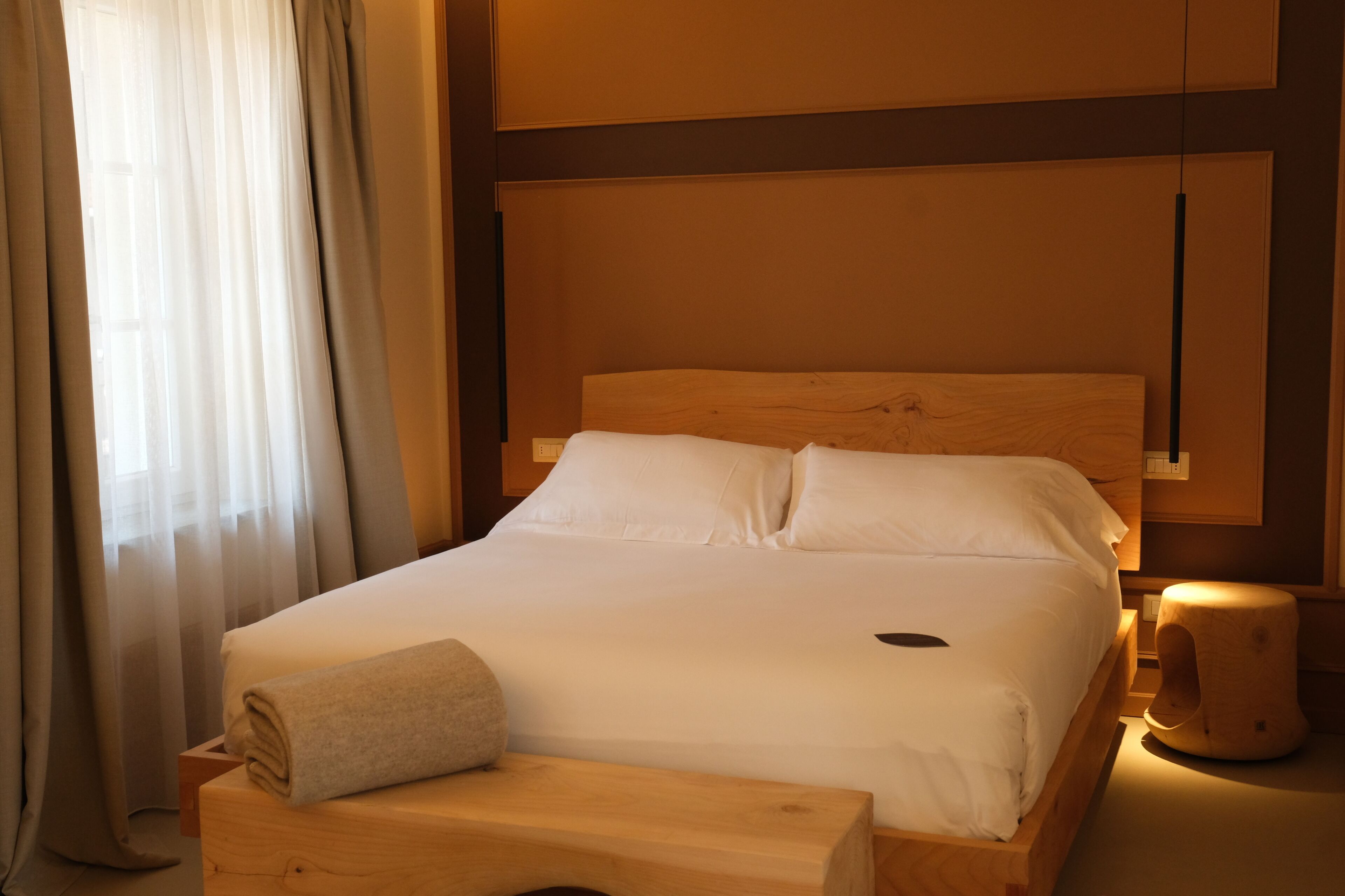 Una cama bien hecha con sábanas blancas, cabecera de madera y mesita de noche con una toalla enrollada.