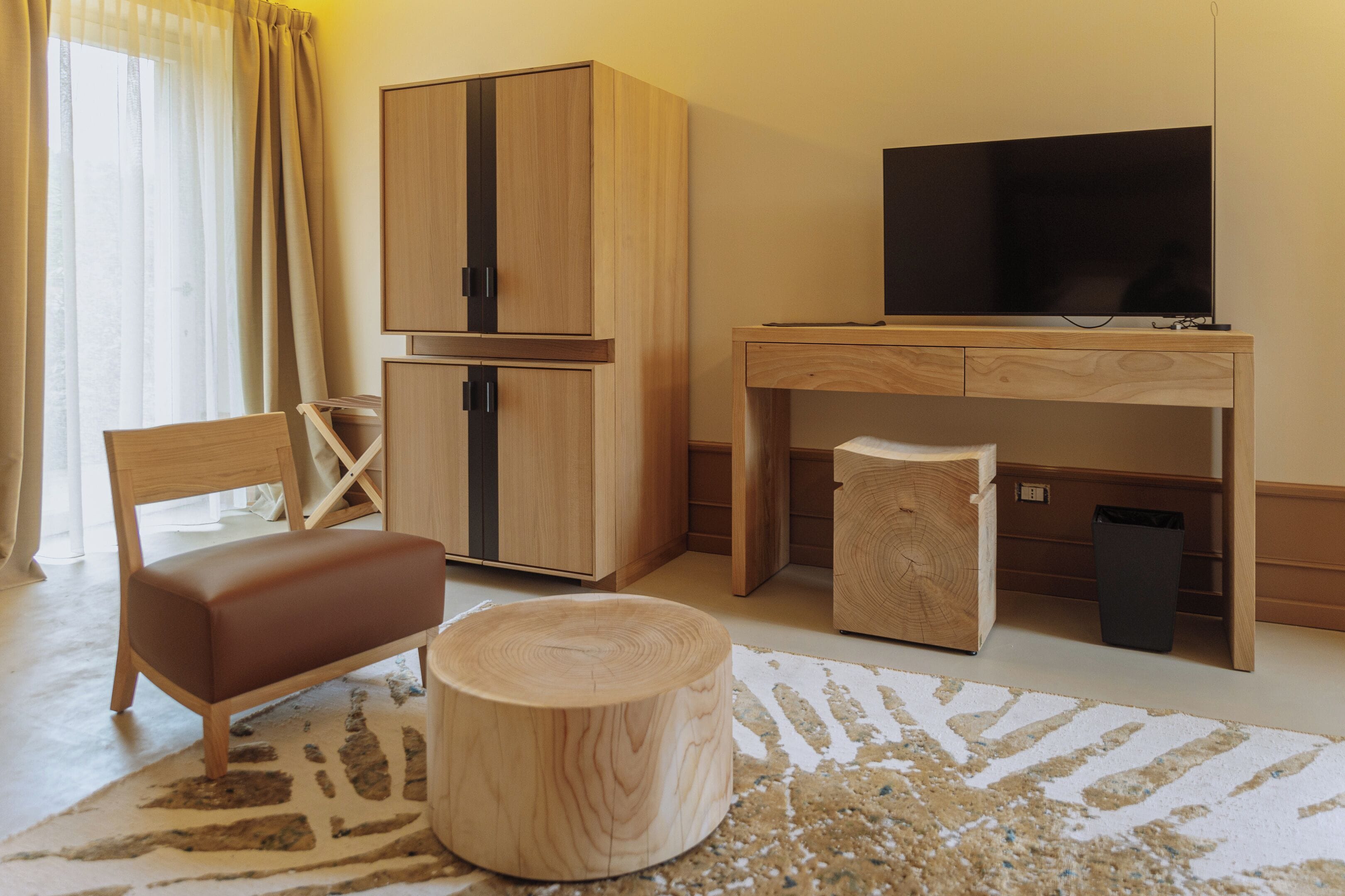 Ein Hotelzimmer mit einem Holztisch, einem Fernseher, einem braunen Ledersessel, einem Holzschemel und einem großen Fenster mit Vorhängen.