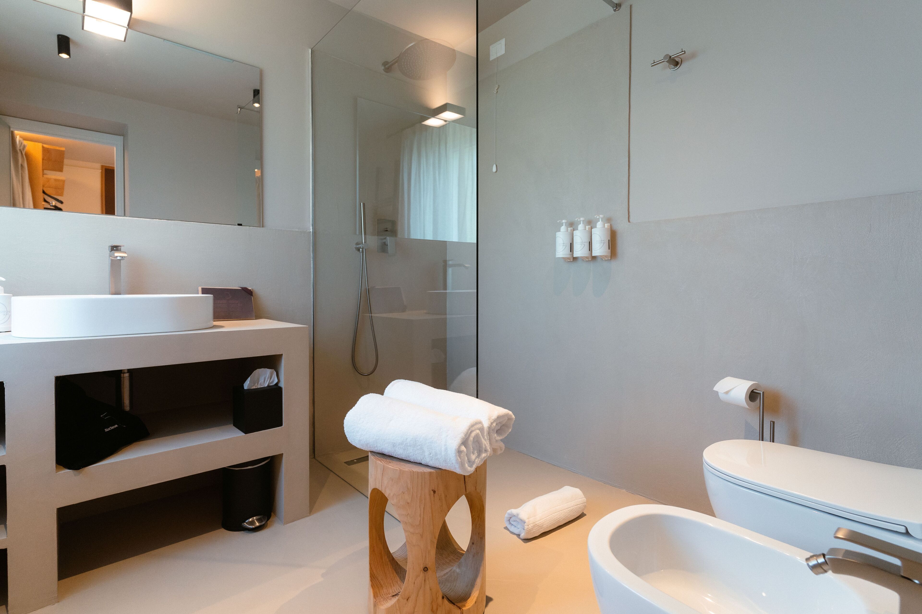 A modern bathroom with a glass shower enclosure, a white sink, and a wooden stool with towels.