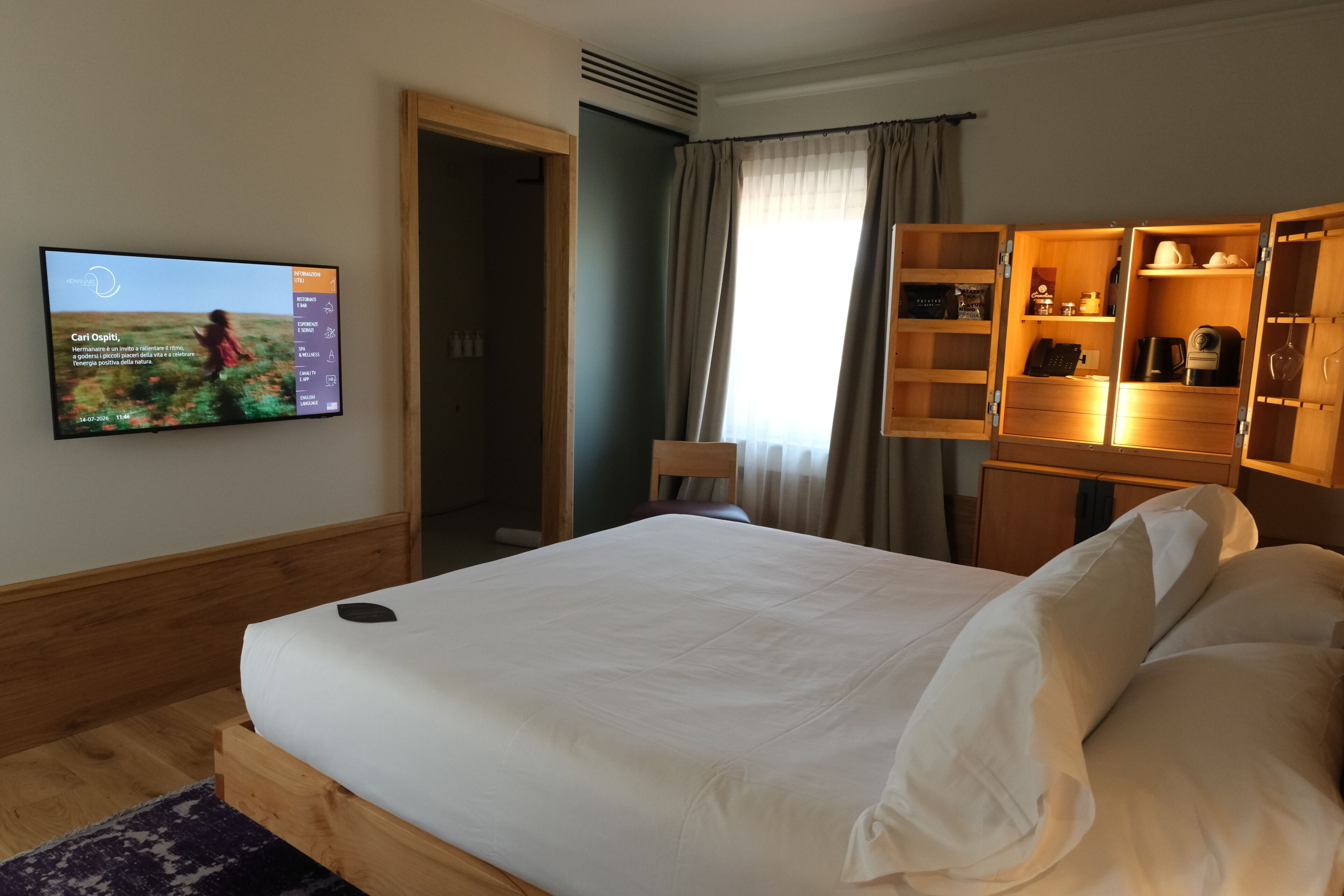 Hotellihuone, jossa on sänky, televisio, jossa näkyy ruokalista, vaatekaappi hyllyineen ja ikkuna, jossa on verhot.