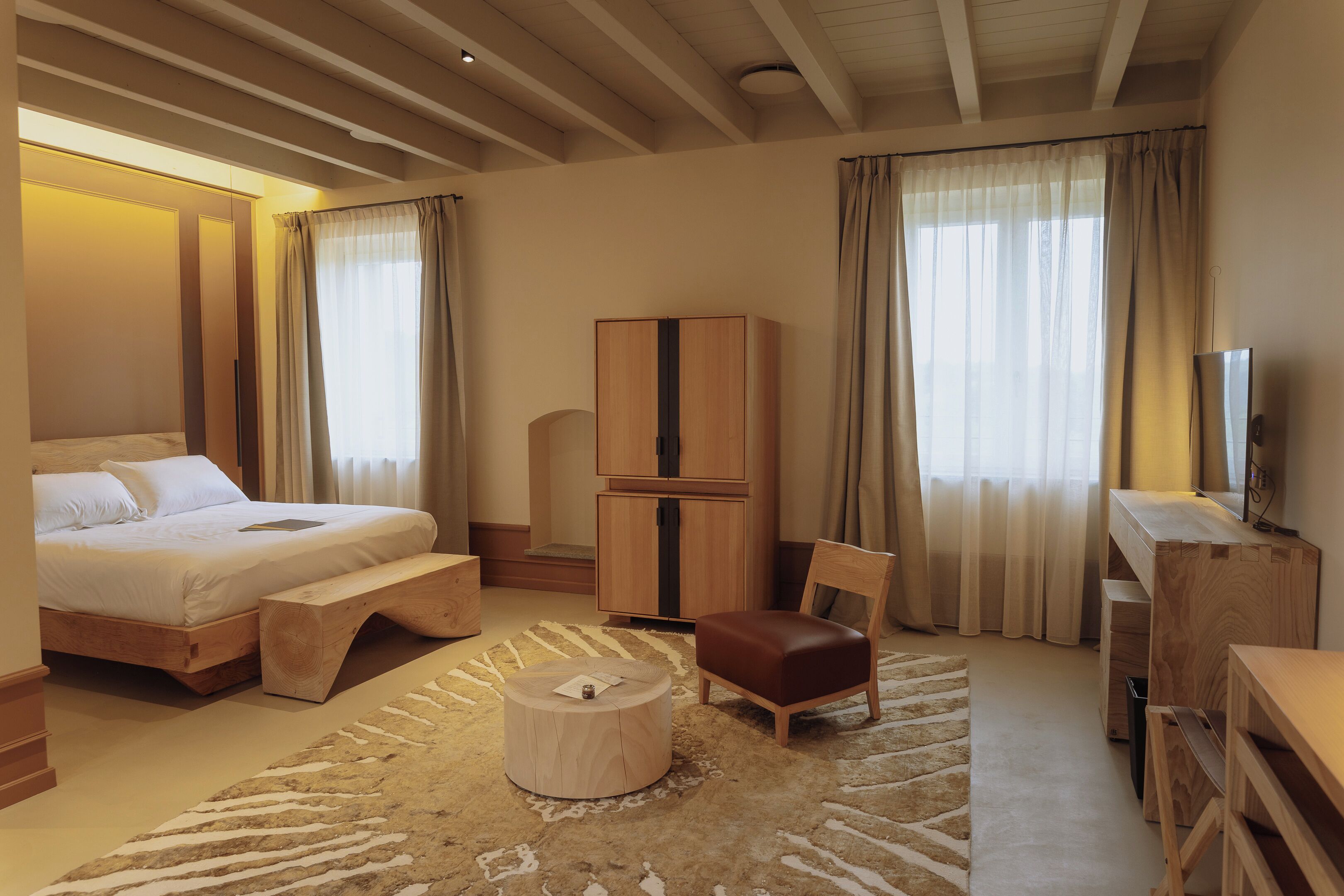 Una habitación de hotel con cama, un banco de madera, una mesa redonda de madera, un armario de madera, un escritorio de madera y un televisor.