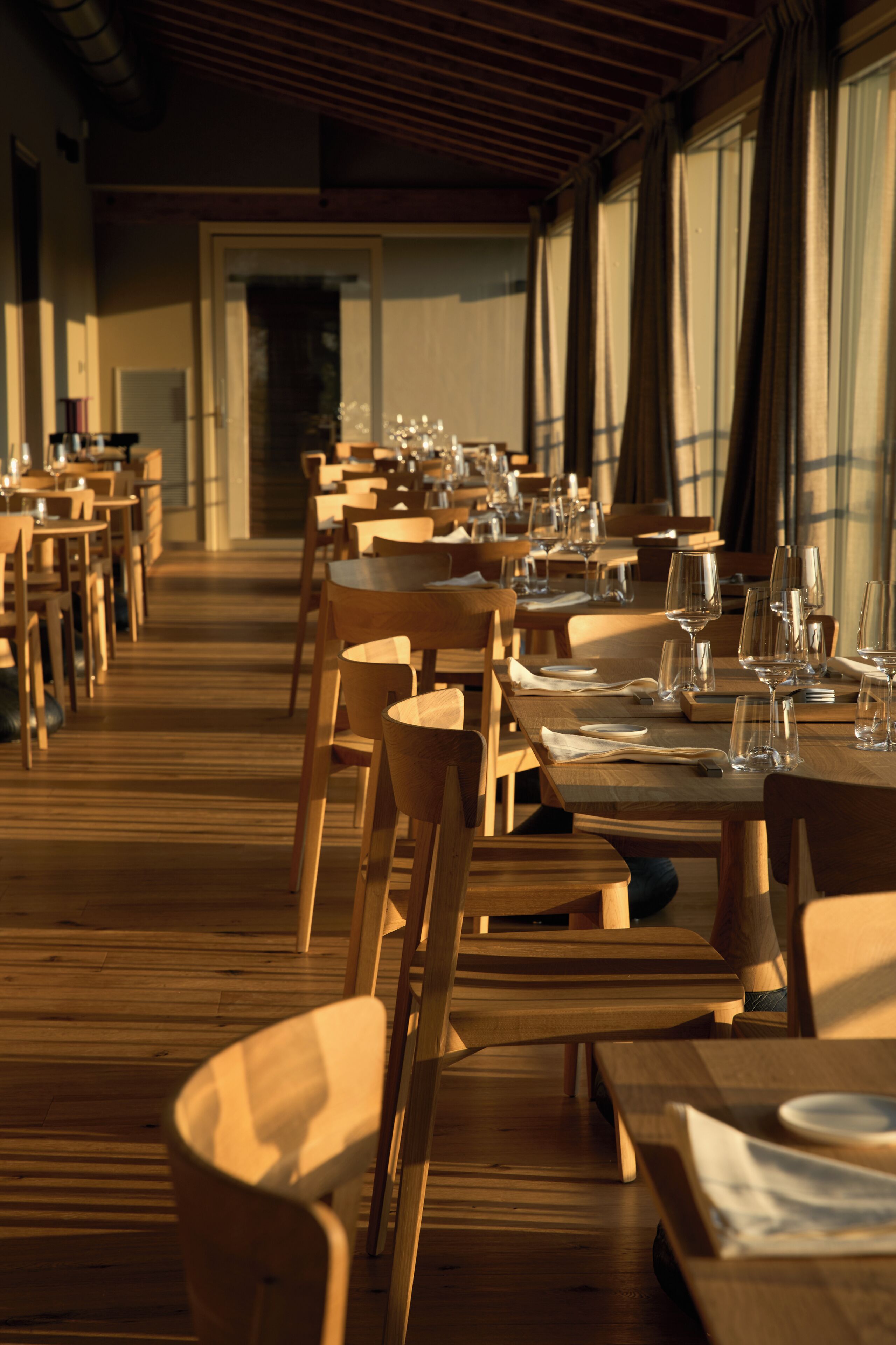 A restaurant with wooden tables and chairs, arranged in rows, with empty wine glasses and plates on the tables.