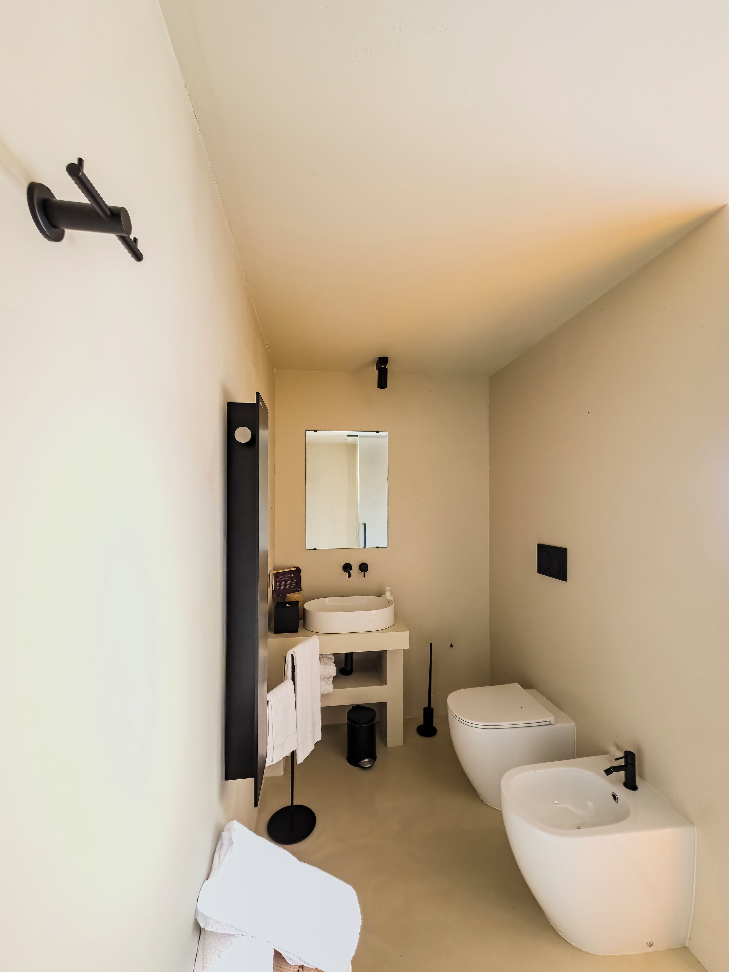 Un baño moderno con un lavamanos blanco, un bidé y un inodoro. Hay un espejo sobre el lavamanos y una toalla colgada en la pared.