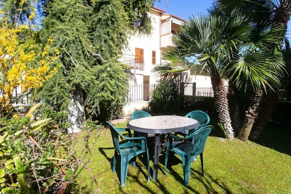 A round table with four chairs in a garden setting, surrounded by greenery and a building in the background.