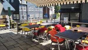 An outdoor seating area with tables and chairs, a covered section, and a view of traditional buildings in the background. - Rennsteighotel Grüner Baum (Suhl)