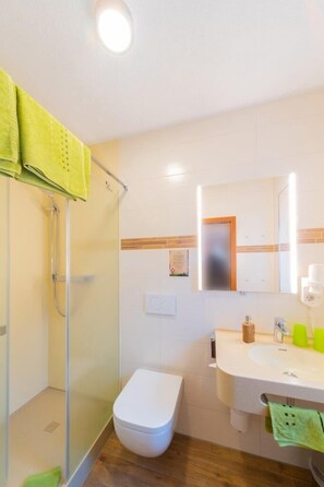 Un baño con cabina de ducha de vidrio, inodoro blanco, lavamanos con espejo y una toalla verde colgada en la pared.