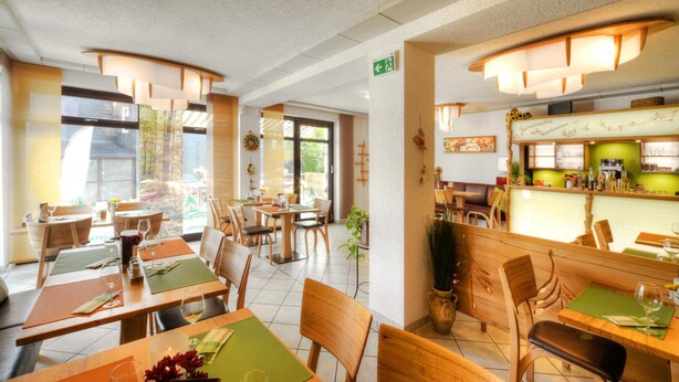 A restaurant with wooden tables and chairs, a bar area, and large windows allowing natural light. - Rennsteighotel Grüner Baum (Suhl)