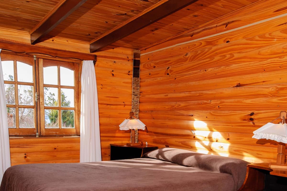 Un dormitorio de cabaña de madera con cama, mesita de noche y ventana con cortinas.