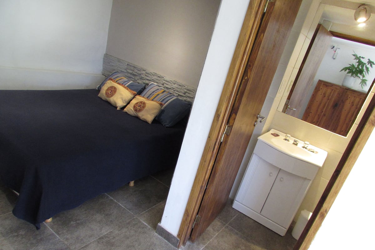 Un dormitorio con cama, almohadas, una mesita de noche y un baño con lavabo y espejo.