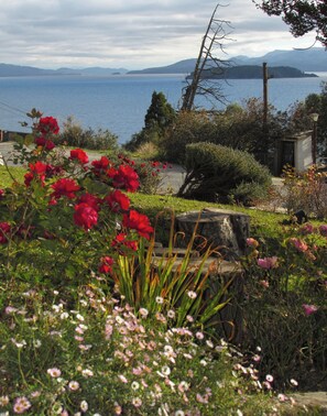 Ein Garten mit verschiedenen Blumen und Blick auf ein Gewässer, in der Ferne Berge.