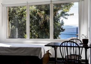 Une pièce comprenant un lit, une table et une chaise. Une grande fenêtre offre une vue sur des arbres et un plan d’eau.