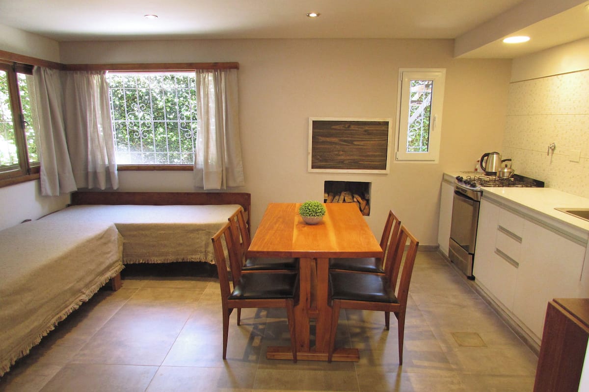Una habitación compacta con una mesa de comedor de madera, cuatro sillas, un banco, una pequeña zona de cocina con estufa y una ventana con cortinas.