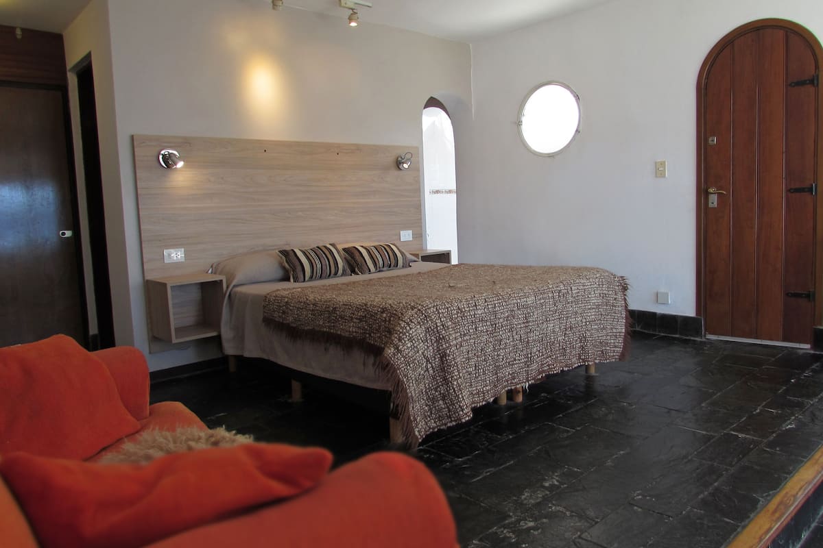 Un dormitorio con cama, mesita de noche, cabecero de madera, espejo redondo, puerta de madera y una silla roja.