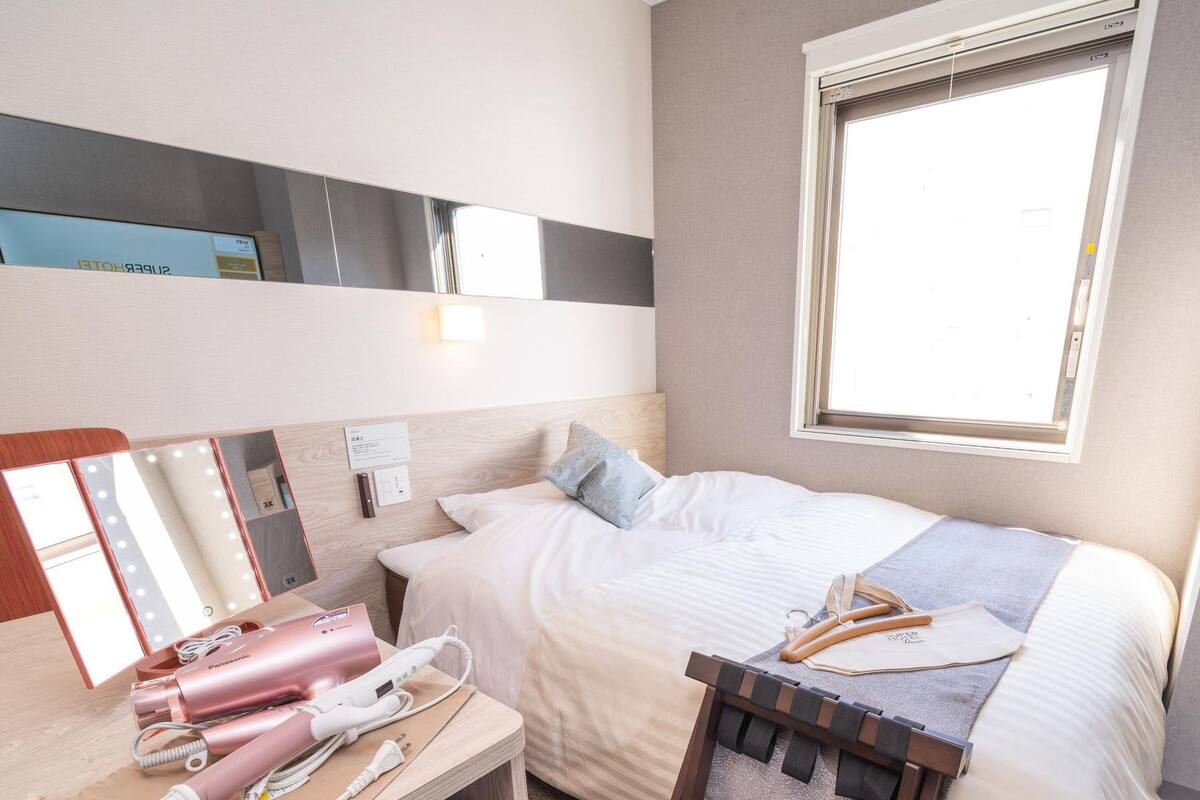 A hotel room with a bed, bedside table, vanity mirror, and a window.