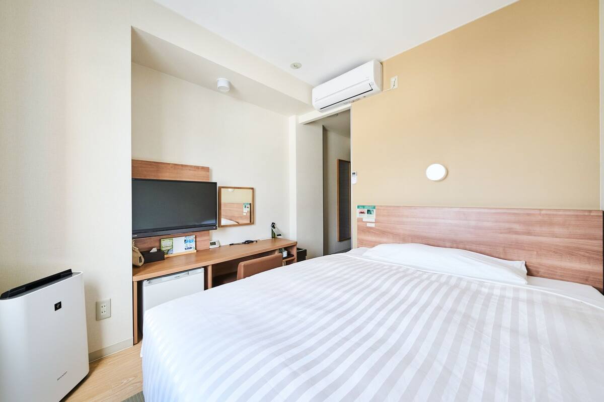 A hotel room with a bed, a desk with a flat-screen TV, a mini-fridge, and an air conditioning unit.