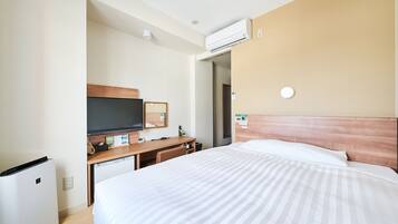 A hotel room with a bed, a desk with a flat-screen TV, a mini-fridge, and an air conditioning unit.