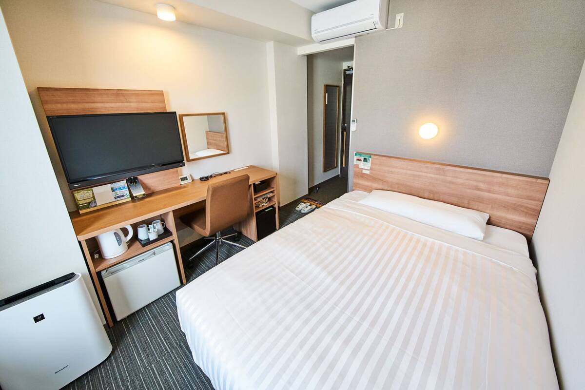 A hotel room with a bed, a desk with a computer, a television, a small refrigerator, and an air conditioning unit.
