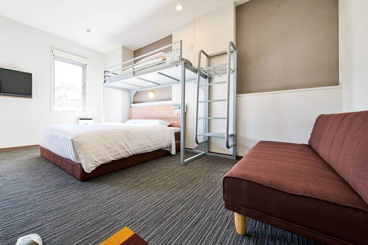 A hotel room with a bunk bed, a sofa, a flat-screen TV, and a window.