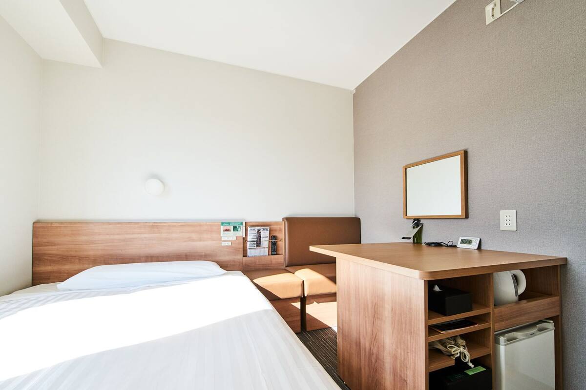 A hotel room with a bed, a desk, a mirror, a small refrigerator, and a wall-mounted light.