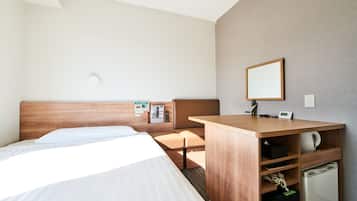 A hotel room with a bed, a desk, a mirror, a small refrigerator, and a wall-mounted light.