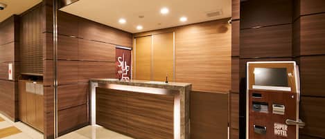 A hotel reception area with a wooden reception desk, a digital display, and a 'Super Hotel' sign on the door.