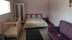 A bedroom with a crib, a bed, a purple sofa, a small table, and a window. - Alex's Studio (Heraklion)
