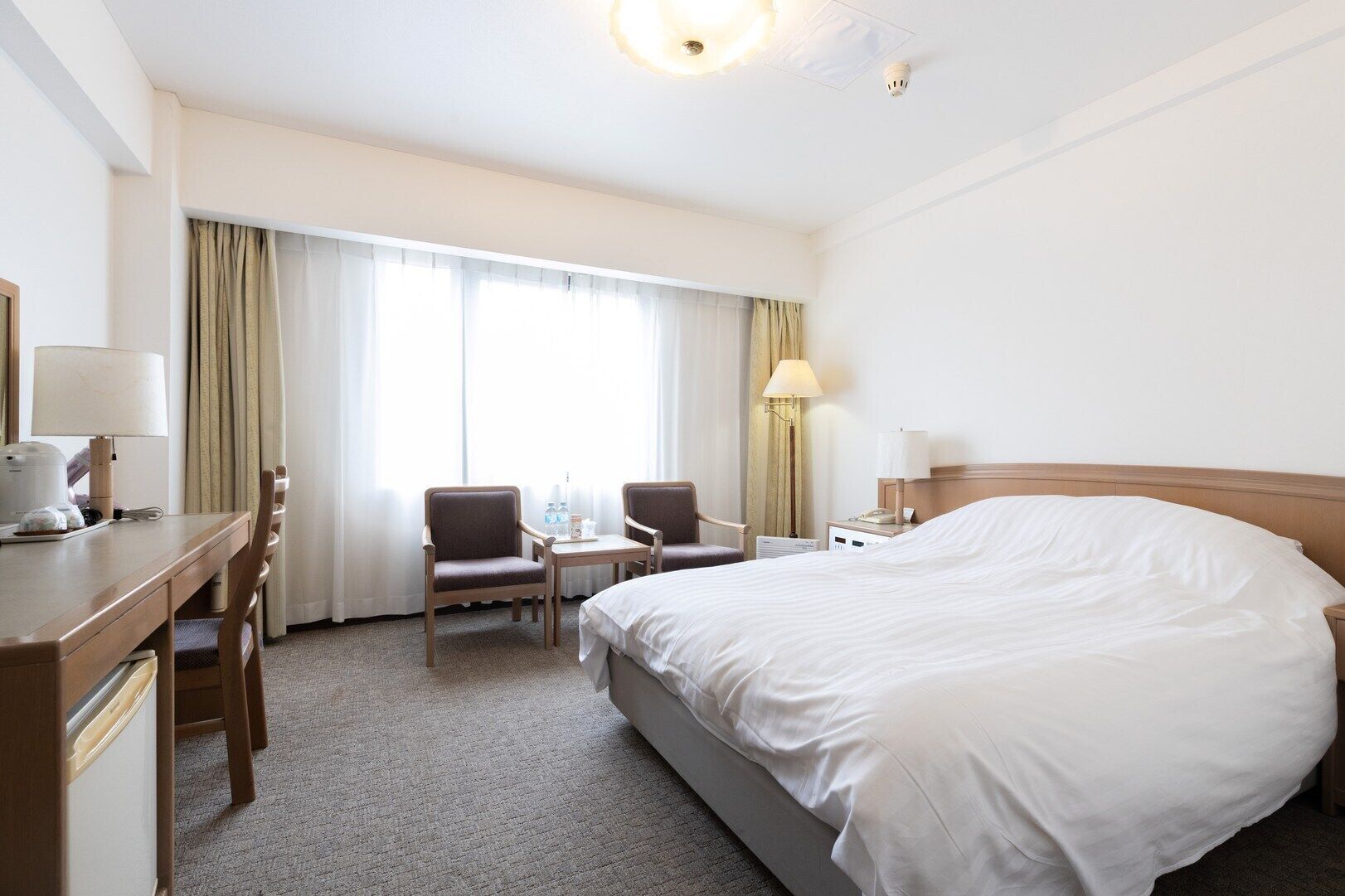 A hotel room with a bed, a desk, two chairs, a table, a lamp, and a window with curtains.