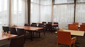 Un salón de conferencias con mesas y sillas, amplias ventanas con cortinas translúcidas y suelo alfombrado.