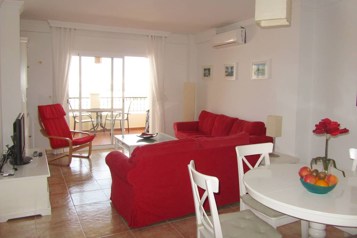 Una sala de estar con un sofá rojo, una mesa de centro blanca y una zona de comedor con una mesa y sillas blancas. Hay un balcón con una silla y una mesa, y un equipo de aire acondicionado en la pared.