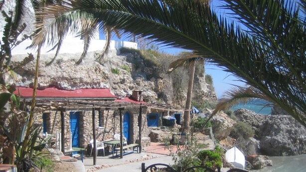 A coastal building with a red roof, stone walls, and a sheltered outdoor seating area with blue doors.