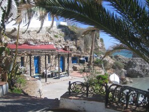 A coastal building with a red roof, stone walls, and a sheltered outdoor seating area with blue doors. - Carabeo 2000 3.1 (Nerja)