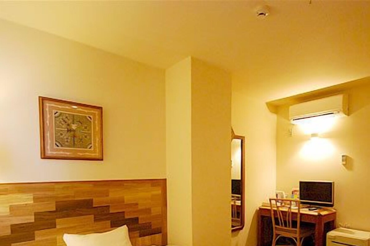 Ett hotellrum med en sänggavel i trä, en säng, ett skrivbord med en stol, en tv och ett litet skåp.
