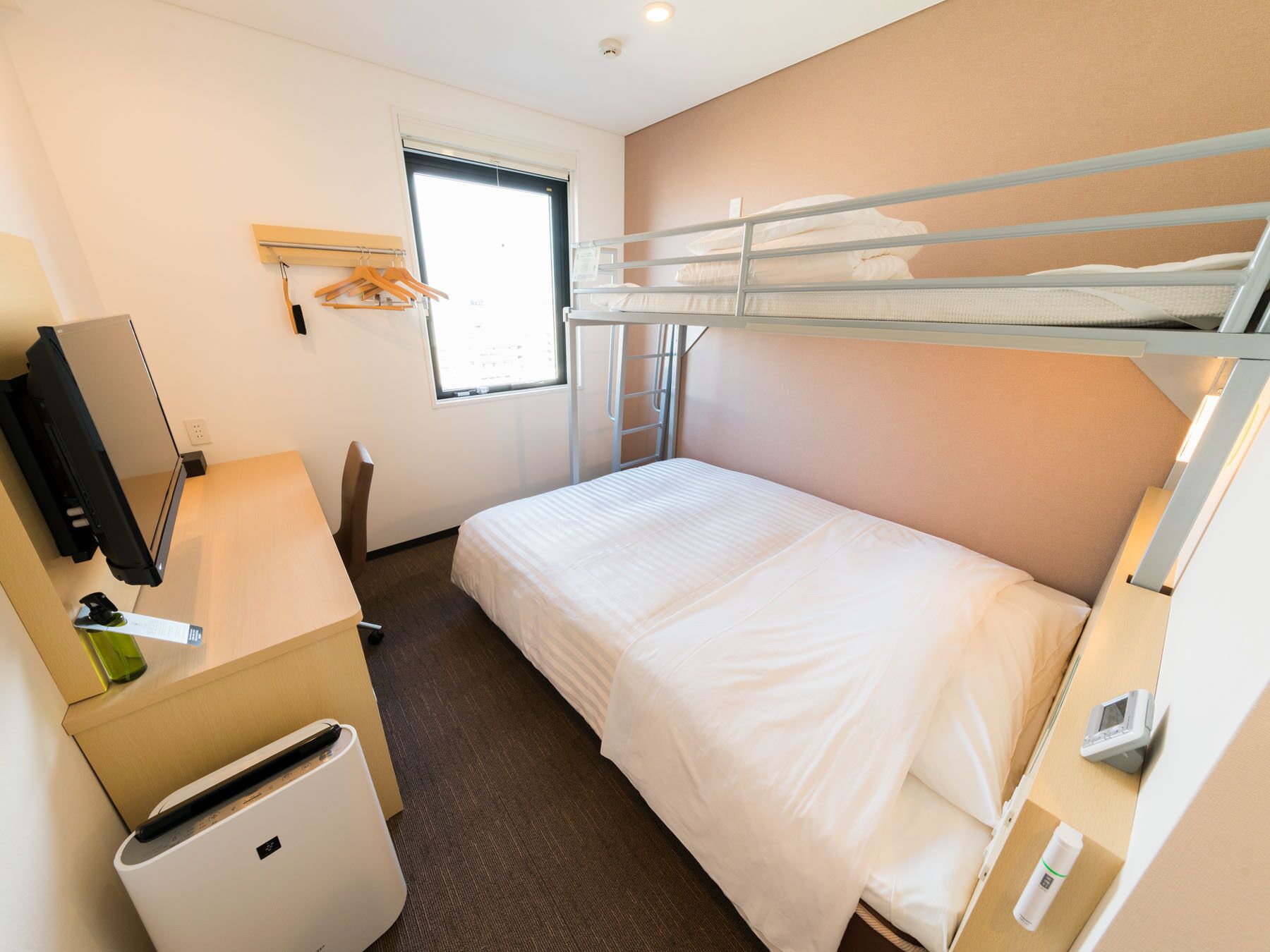 A compact hotel room with a bunk bed, a desk with a computer, a small fridge, and a window with blinds.