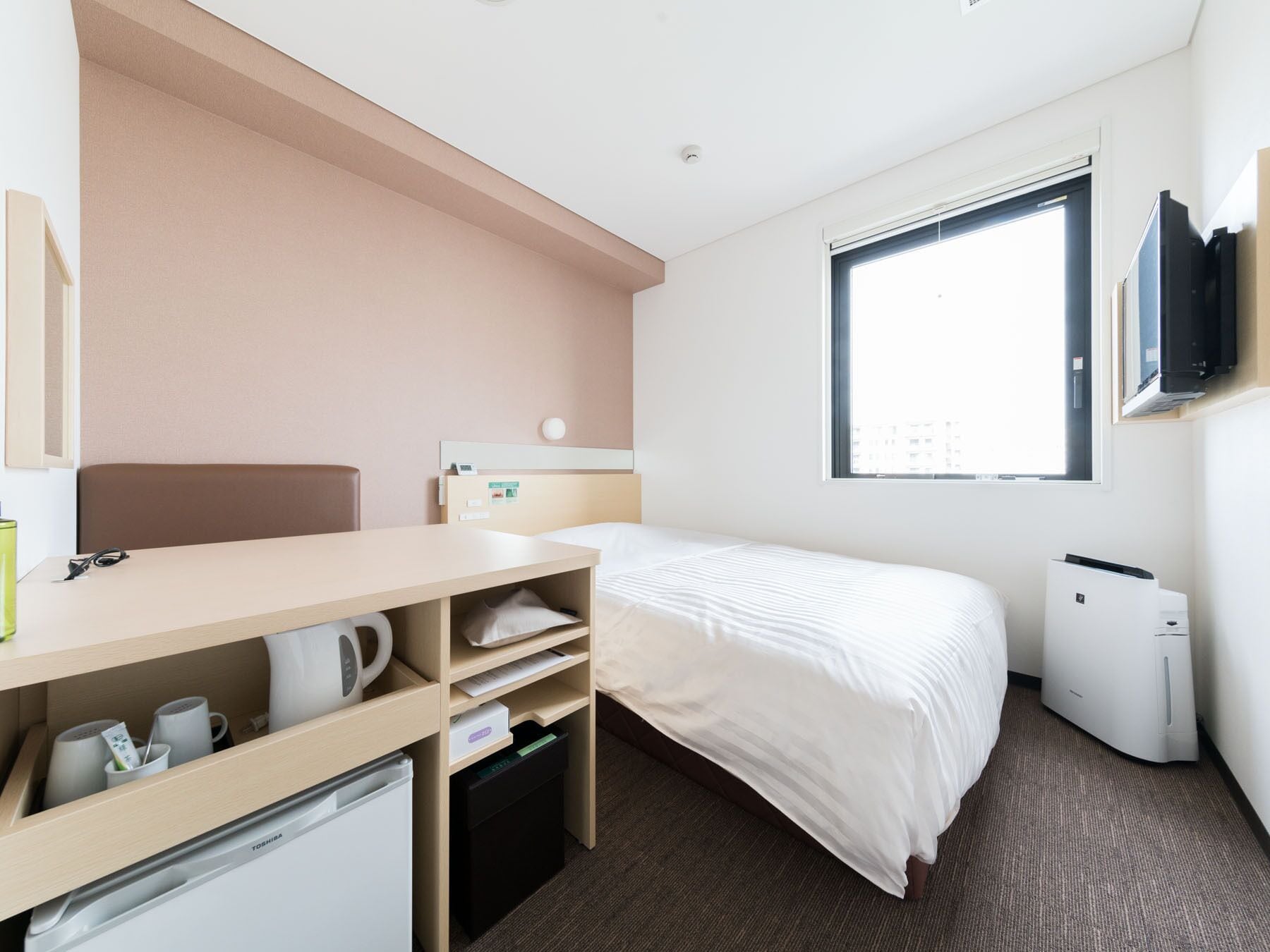 A hotel room with a bed, a desk, a chair, a small refrigerator, a trash bin, and a window with a view of buildings.