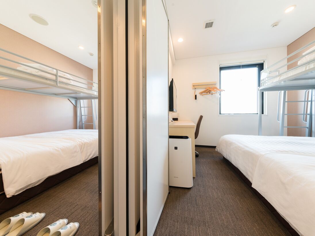 Une chambre d’hôtel avec des lits superposés, un bureau, une chaise, un petit radiateur et une fenêtre.