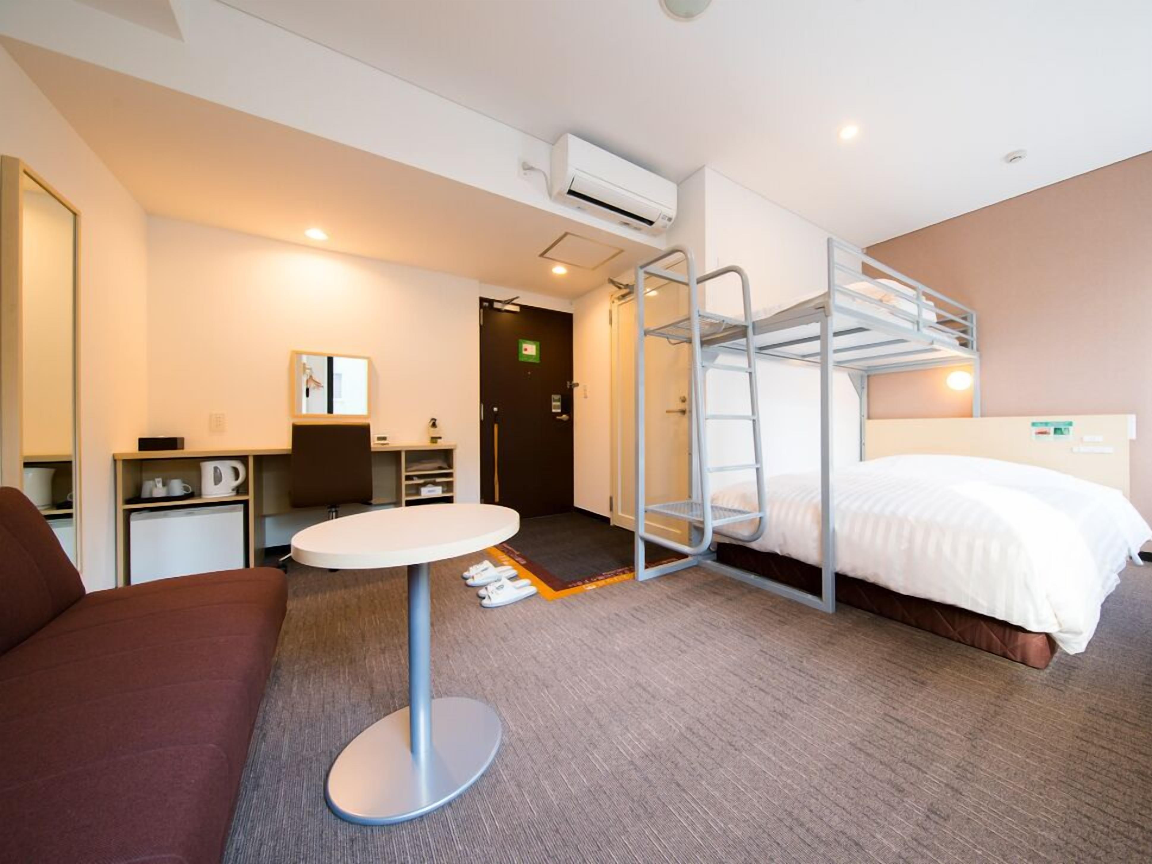 Une chambre d’hôtel avec des lits superposés, une table ronde, un canapé, un bureau avec une chaise et une machine à café.