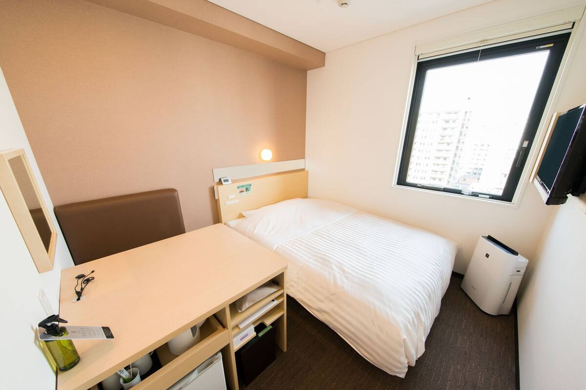 Kompakti hotellihuone, jossa on yksi sänky, työpöytä tuolilla, pieni jääkaappi, roskakori ja ikkuna, josta on näkymä rakennuksiin.