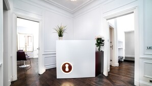 Uma área de recepção com design moderno, destacando um balcão branco com um logotipo circular, uma planta em vaso e um vaso com flores.