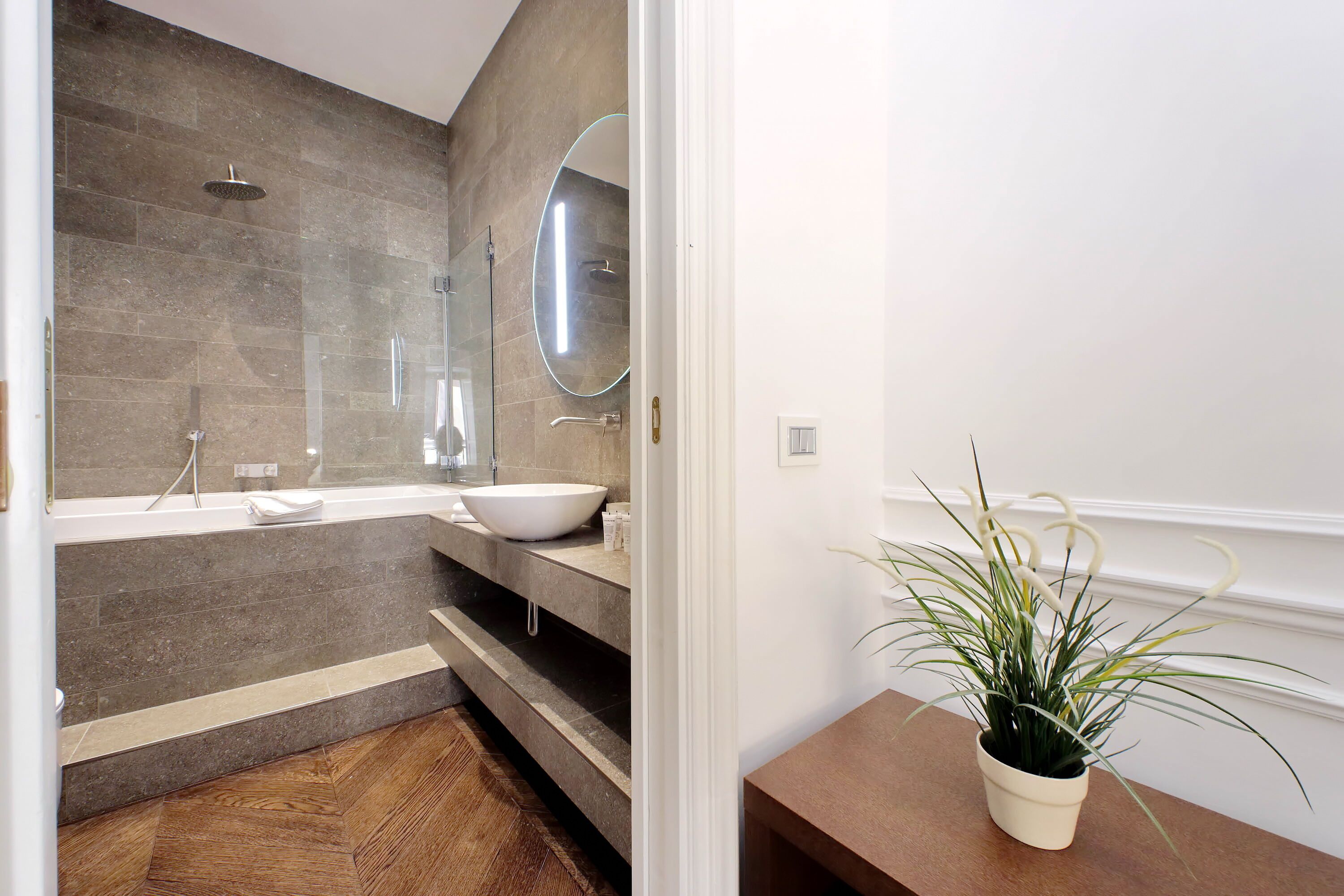 Un bany modern amb una banyera gran, un lavabo i un mirall. El terra és de fusta i hi ha una planta en test sobre una taula de fusta.