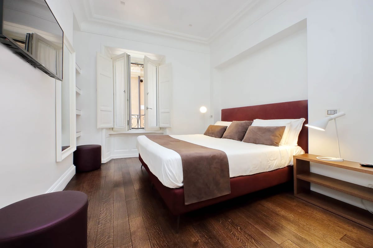 A modern bedroom with a large bed, a bedside table with a lamp, a window with white shutters, and a small purple ottoman.