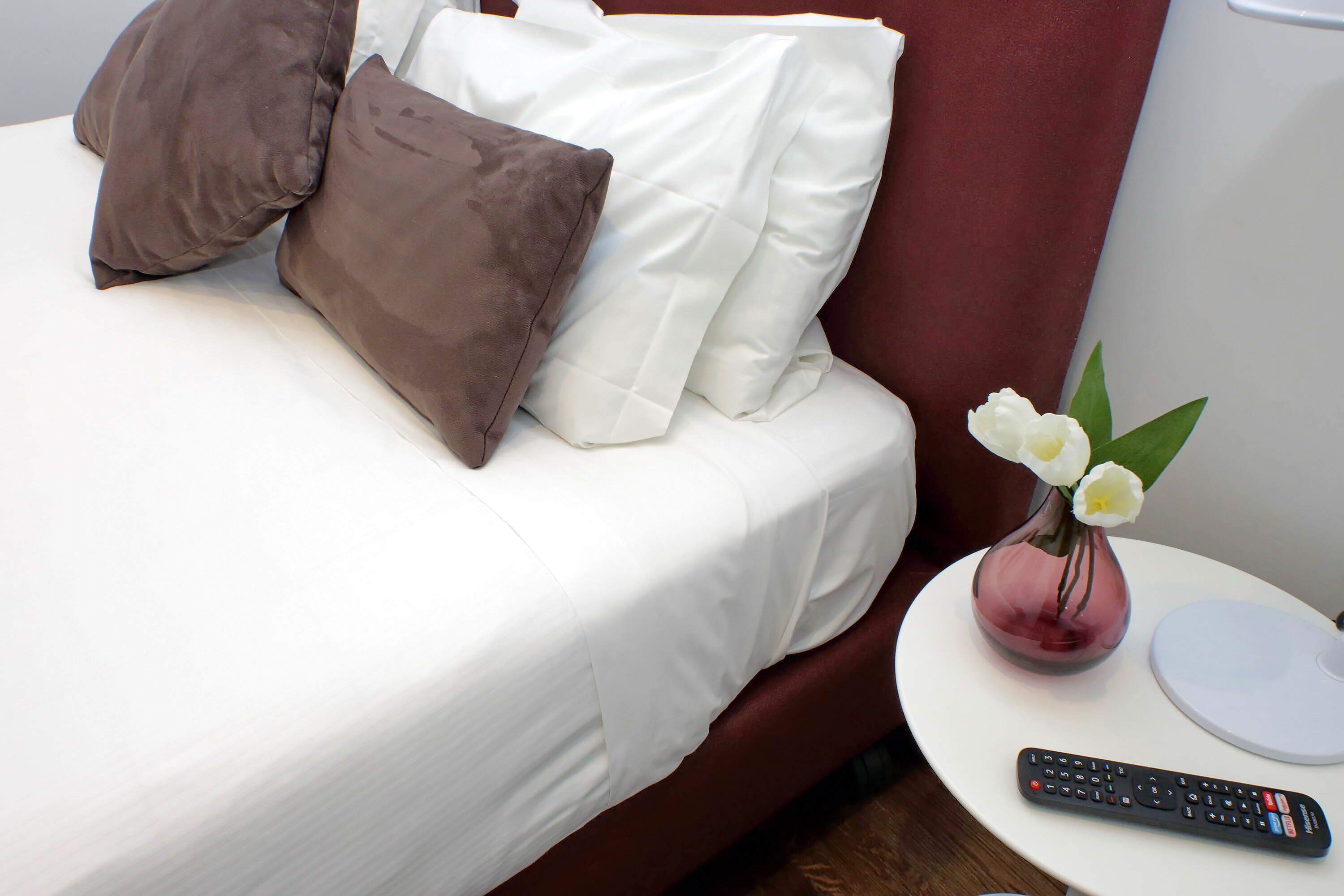 A neatly made bed with white and brown pillows, a remote control, and a vase with white flowers on a small table beside the bed.