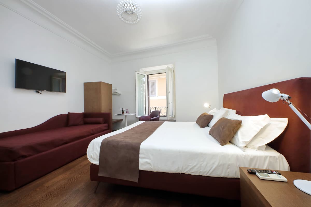 A hotel room with a large bed, a red sofa, a TV mounted on the wall, a desk with a lamp, and a view of the outside balcony.