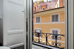 Vista a través de una ventana con una puerta blanca, mostrando un balcón con barandilla de hierro forjado y un edificio amarillo con contraventanas.